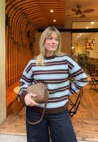 Femme aux cheveux blonds portant un pull rayé marron et bleu clair, tenant un sac à main de créateur marron, debout à l'intérieur d'un café avec une décoration en bois.