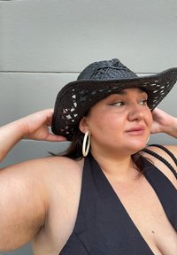 Schwarzer geflochtener Strohhut mit breiter Krempe und offenem Muster. Große Creolen. Dunkelblaues Haltertop, minimale Textur. Neutrale Hintergrund.