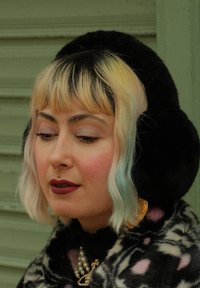 Svarte faux fur øreklokker rammer inn ansiktet til en person med pastellblondt hår med blå tupper. De har på seg dristig leppestift og utsøkte øredobber.