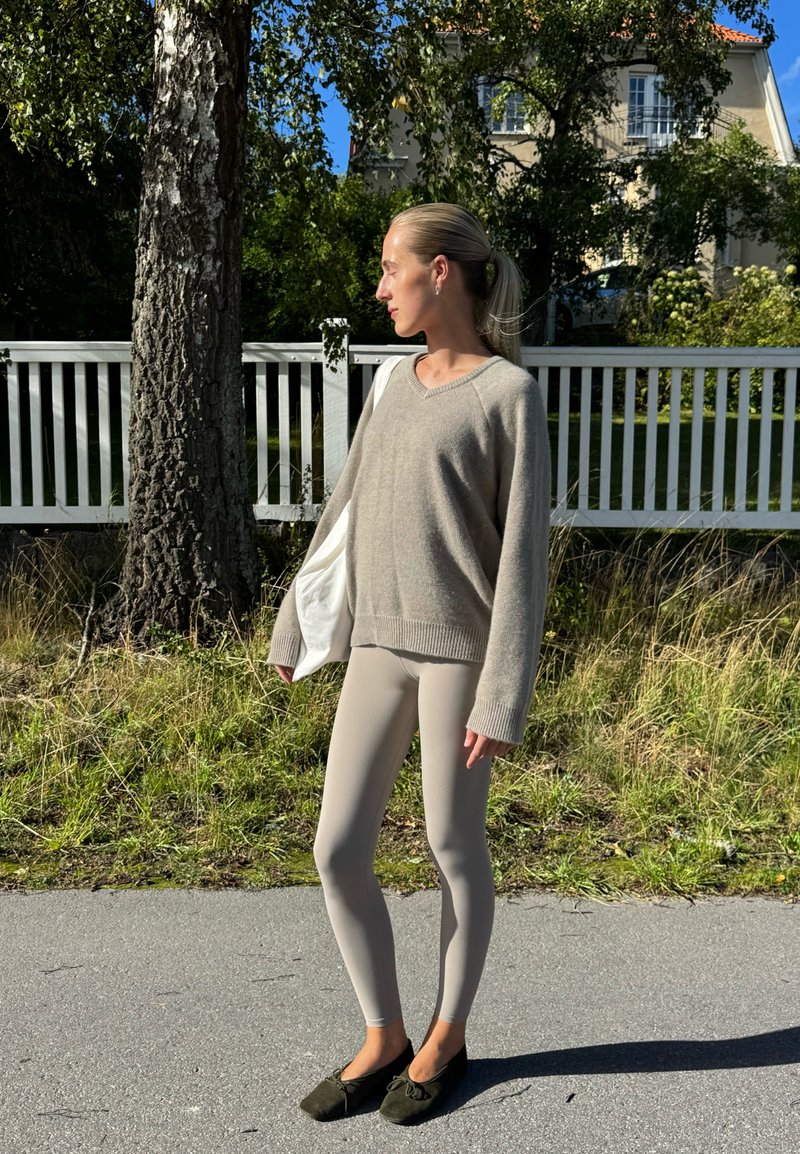 Maglione beige lavorato a maglia con maniche ampie, abbinato a leggings beige chiaro e scarpe slip-on verde scuro, in piedi su un sentiero pavimentato.