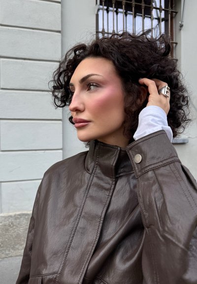 Chaqueta de cuero marrón con un cuello alto y botones de presión, con una textura suave y un brillo sutil. El modelo tiene el cabello rizado y lleva un anillo de plata.