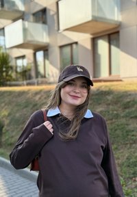 Jonge vrouw in een lange mouwen bruin shirt en een vos cap staat glimlachend buiten bij een modern appartementencomplex, terwijl ze de schoudertas vasthoudt aan de strap.