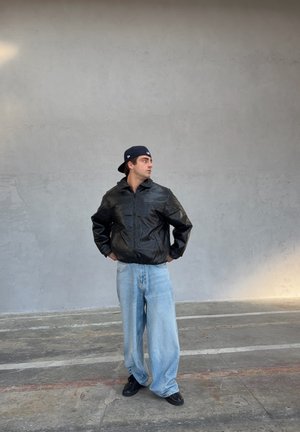 Chaqueta bomber de cuero negro con cierre de cremallera, combinada con jeans sueltos de mezclilla azul claro. Zapatillas negras completan el conjunto.