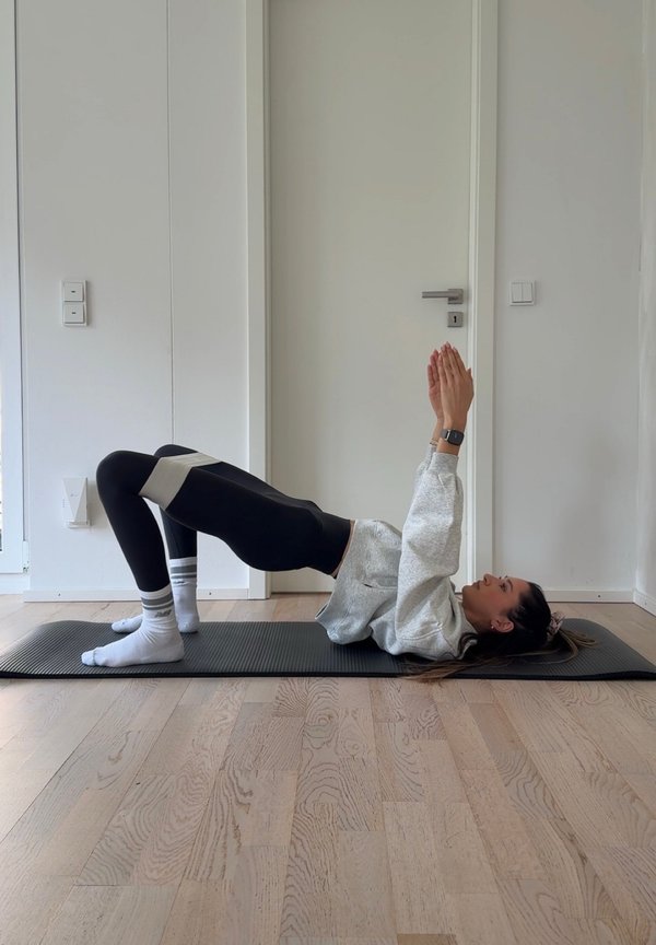 Henkilö jumppamatolla tekee pakarasiltaa vastuskuminauha polvien yläpuolella, kädet ojennettuna ylöspäin, pienessä huoneessa, jossa valkoiset seinät ja puulattia.