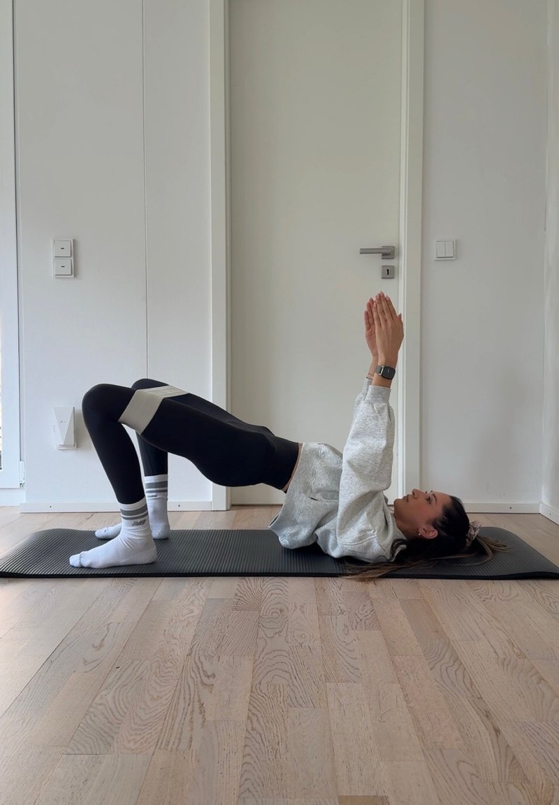 Άτομο σε στρώμα γυμναστικής εκτελεί γέφυρα γλουτού με λάστιχο αντίστασης πάνω από τα γόνατα, τα χέρια τεντωμένα προς τα πάνω, σε έναν μικρό χώρο με λευκούς τοίχους και ξύλινο πάτωμα.