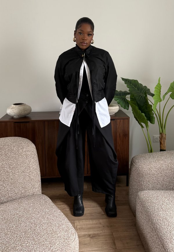 Junge Frau steht zwischen zwei beigen Sesseln im Wohnzimmer, trägt ein schwarz-weißes geschichtetes Outfit mit weitem Hosenbein, schwarzen Stiefeln und goldenen Creolen.