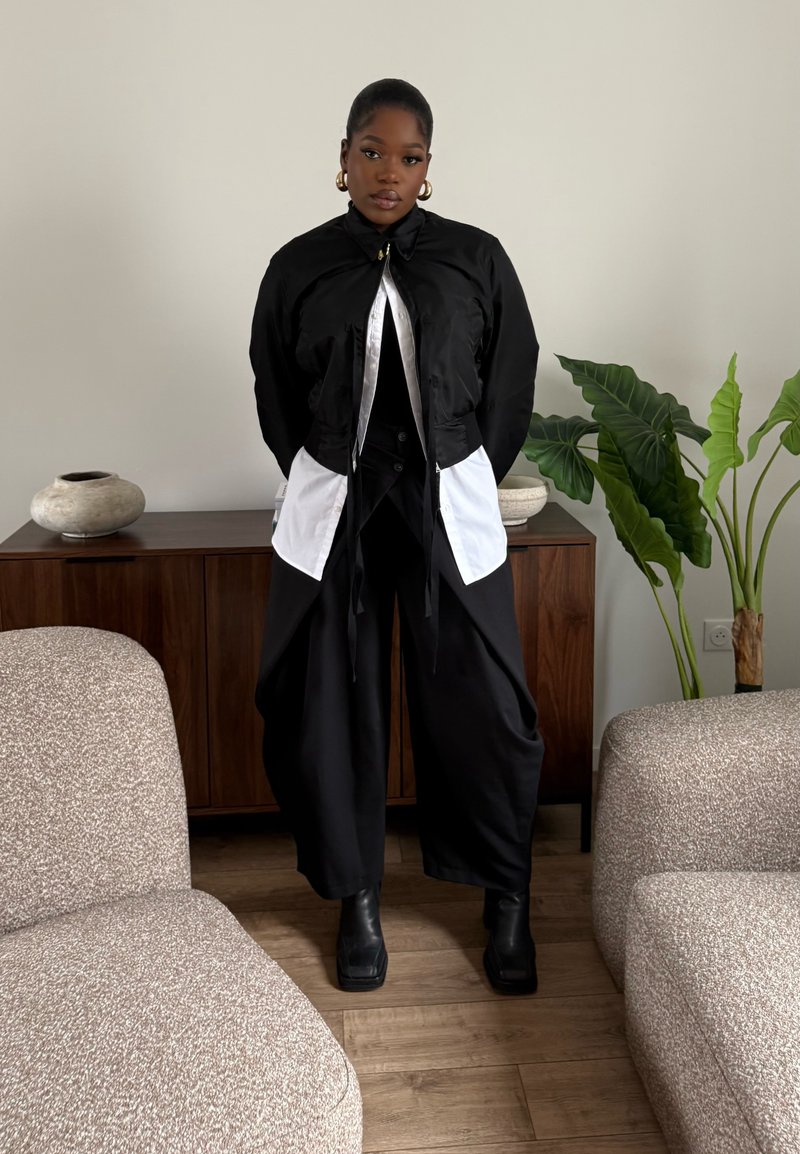Jeune femme debout entre deux fauteuils beiges dans un salon, portant une tenue superposée noir et blanc avec un pantalon large, des bottes noires et des boucles d'oreilles créoles dorées.