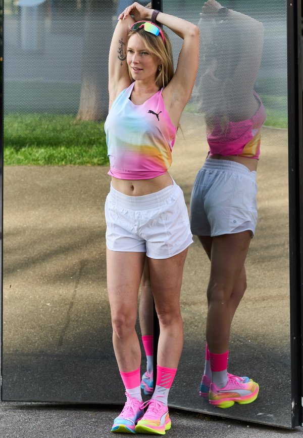 Kvinde iført multifarvet sportstop, hvide shorts og lyse sneakers strækker sig med armene hævet foran en reflekterende overflade udendørs.