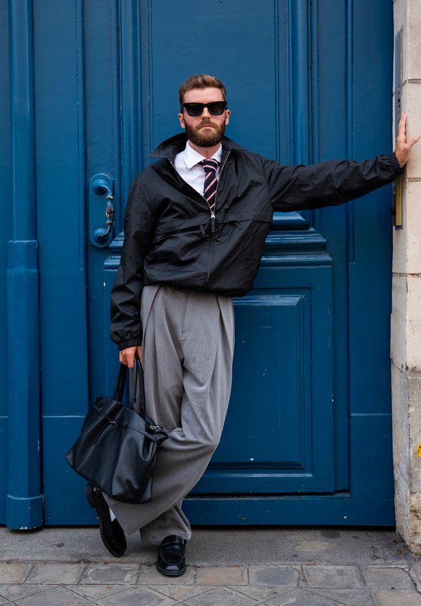 Man in sunglasses and black jacket leaning against blue doorframe holding black bag, wearing grey trousers and black shoes.