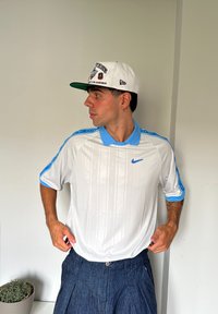 Balta Nike polo marškiniai su šviesiai mėlynomis apykaklės ir rankovių detalėmis; turi subtilų vertikalų prekės ženklo ženklinimą. Nešiojama su mėlynomis džinsinėmis šortais ir balta kepure.