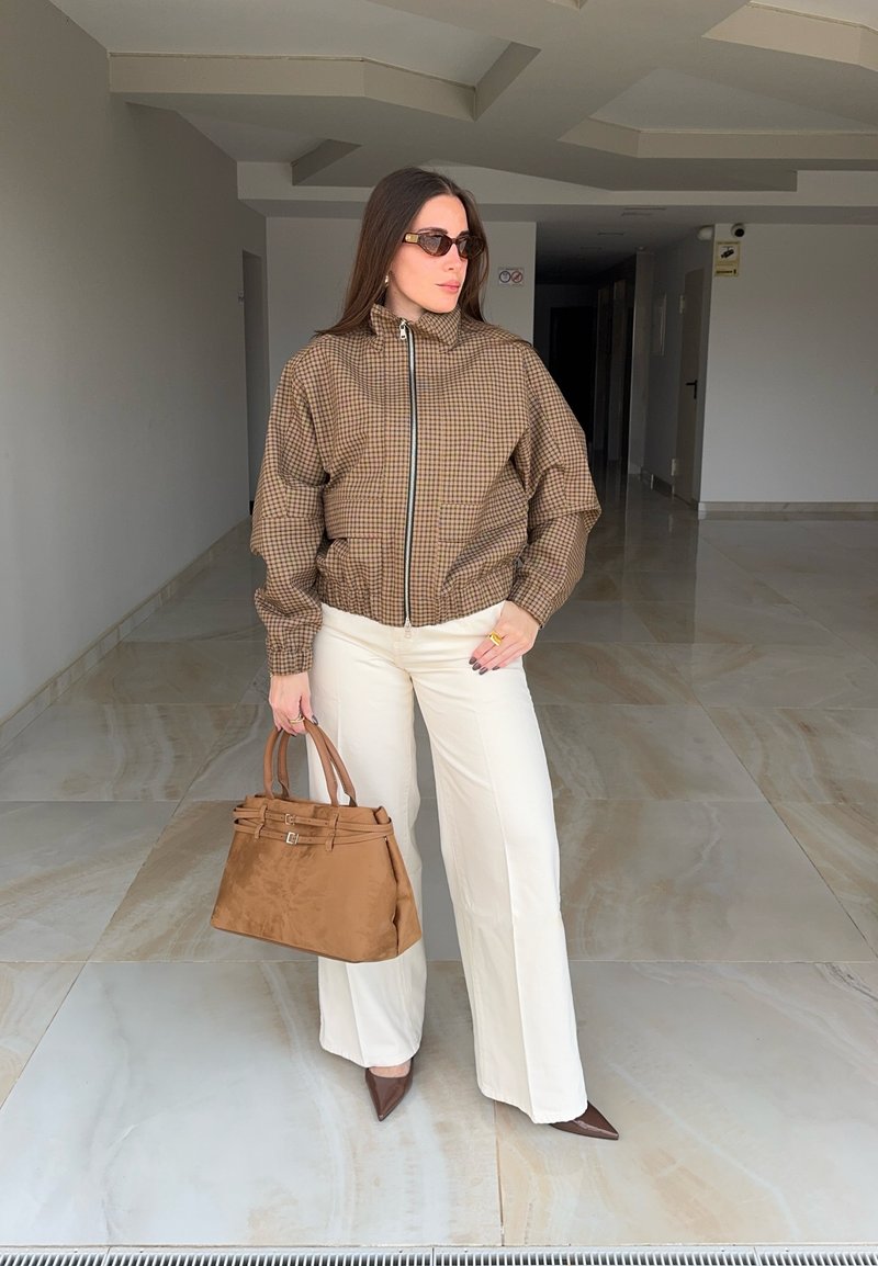 Femme portant une veste marron à carreaux, pantalon blanc, chaussures marron pointues, lunettes de soleil, tenant un grand sac à main marron à l'intérieur.