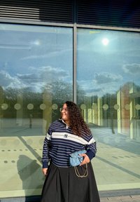 Mujer con cabello rizado y gafas sonríe, vistiendo un suéter a rayas en tonos marino y blanco y una falda negra, sosteniendo un bolso azul frente a una pared de cristal.