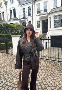 Femme portant une veste en cuir noire ceinturée et un chapeau marron, tenant un sac marron, debout sur des pavés devant des maisons mitoyennes blanches.