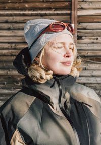 Veste isolante en noir, gris et beige ; dotée d'un col montant et d'une fermeture éclair. Portée avec un bonnet gris et des lunettes de soleil sur le front.
