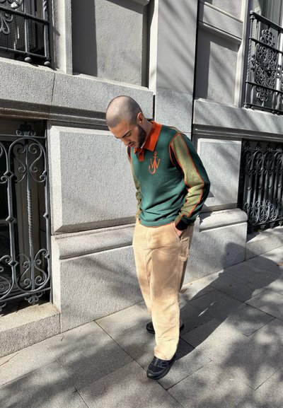 Polo verde y naranja con cuello, que presenta un logo de ancla; combinado con pantalones de pana beige y zapatos negros. Fondo urbano iluminado por el sol.