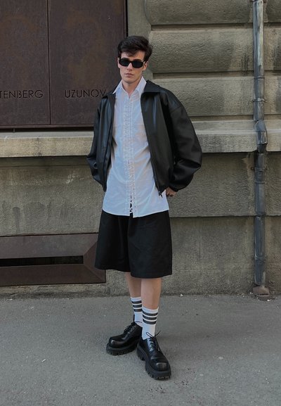 Chaqueta de cuero negra sobre una camisa blanca de botones, pantalones cortos negros y zapatos negros gruesos, combinados con calcetas blancas a rayas.