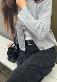 Cardigã cinza com nervuras e fecho de botão, usado sobre uma blusa branca. Combinado com calças cargo pretas com bolsos laterais e uma mala preta.