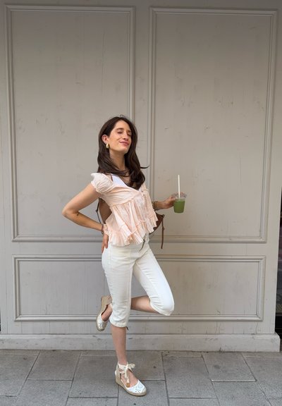Mujer con pantalones blancos cortos y top melocotón con volantes, de pie sobre una pierna, sosteniendo una bebida verde, con los ojos cerrados y sonriendo contra una pared gris pálida.