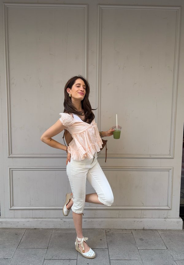 Femeie în pantaloni albi scurți și top piersică cu volane care stă pe un picior, ținând o băutură verde cu ochii închiși și zâmbind în fața unui perete gri pal.