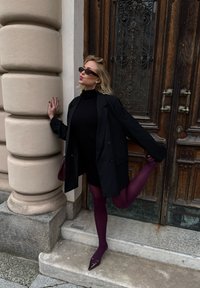 Black oversized blazer over a fitted black dress, paired with burgundy tights and pointed-toe shoes. Background features wooden doors and stone steps.
