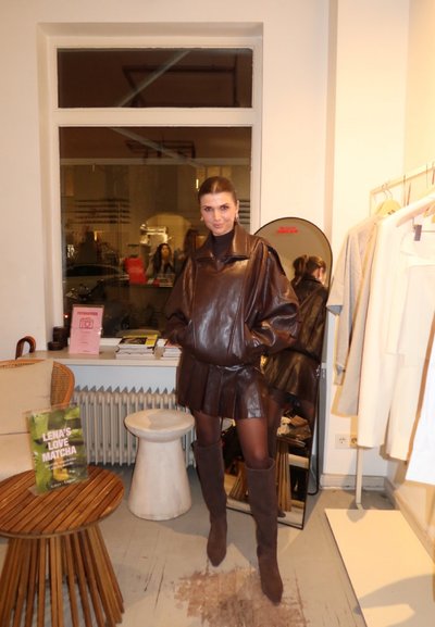 Mujer posando en el interior con una chaqueta de cuero marrón de gran tamaño, falda plisada de cuero y botas hasta la rodilla, junto a un espejo y un perchero.