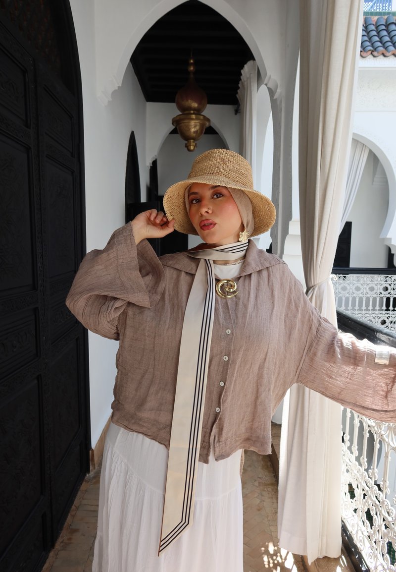 Jonge vrouw die een beige hoed, lichtbruine blouse en een witte rok draagt, staat in een witte gewelfde gang met decoratieve balustrade en gordijnen.