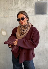 Νεαρή γυναίκα με σκούρα γυαλιά ηλίου φοράει καφέ μπουφάν και μαντίλι με σχέδια, κρατώντας σκοτεινό κόκκινο πλεκτό φάκελο μπροστά σε φόντο από τσιμεντένιο τοίχο.