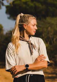 Beige, oversized T-skjorte med rund hals, lagd med et brunt skjerf knyttet rundt halsen. Sorte bukser med snøring. Sirkulær smartklokke på håndleddet.