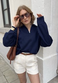 Mörkblå zip-neck sweatshirt med ballongsärmar, krämfärgade shorts, brunt läderbälte och väska samt solglasögon, mot en vit vägg.