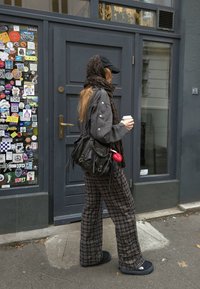 Sieviete pelēkā pārāk lielā krekla ar pogātām piedurknēm, pūkainās biksēs, nēsā melnu somu, tur kafijas krūzi, pastaigā gar durvīm ar uzlīmēm.