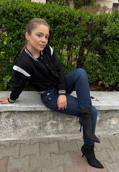 Chaqueta bomber negra con rayas blancas, vaqueros azules desgastados y botas de tacón negro. Sentada en un muro de piedra con follaje verde detrás.