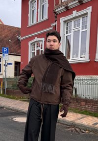 Ruda izoliuota striukė su elastinėmis rankogaliais, derinama su tamsiai rudu šaliku ir pirštinėmis. Tamsios kelnės papildo ansamblį prie plytų fono.