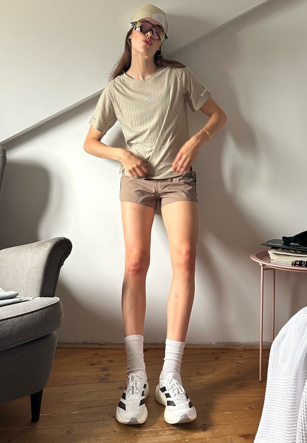 Frau trägt beigefarbenes Outfit, weiße Turnschuhe, Socken, Visier und große Sonnenbrille und steht drinnen in der Nähe eines Sessels und eines runden Beistelltisches.