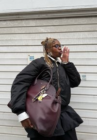 Jachetă bomber neagră, cămașă albă cu guler, fustă neagră, ținând o geantă mare din piele bordo cu accente de breloc, suprafață texturată vizibilă.