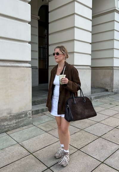 Chaqueta de ante marrón, vestido blanco, zapatillas grises. Bolso negro con detalles claros. Gafas de sol con detalles dorados. Sosteniendo una bebida con pajita.