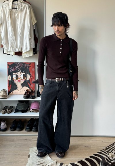 Joven con tatuajes que lleva una camiseta de polo de manga larga de color oscuro y acanalada, vaqueros anchos negros y gafas de sol, con el cabello negro, de pie en el interior cerca de ropa y zapatos.