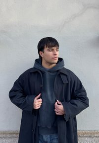 Νέος άντρας που φοράει μαύρο παλτό πάνω από σκούρα κουκούλα, στέκεται μπροστά σε ανοιχτό γκρι τοιχο με υφή, κοιτάζοντας προς τα πλάγια με τα χέρια να διορθώνουν το παλτό του.