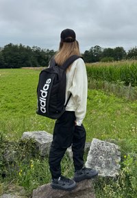 Zaino Adidas nero con logo bianco, indossato sopra una felpa chiara e pantaloni cargo neri. Sneakers grigie completano l'outfit.