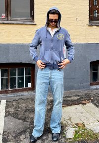 Felpa blu con zip, tasca frontale e logo, abbinata a jeans dritti blu chiaro e scarpe nere. Sfondo urbano con finestre.