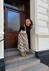 Egy faajtó üvegpanellel és 17-es számmal. Egy személy barna hosszú ujjú felsőben, kockás szoknyában és bézs cipőben áll a kőlépcsőkön.