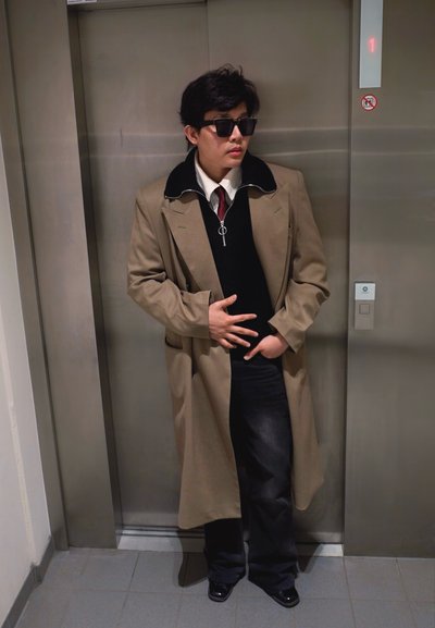 Persona con gafas de sol, abrigo largo beige, pantalones negros y camisa blanca con corbata burdeos, de pie frente a las puertas cerradas del ascensor que muestran el piso 1.