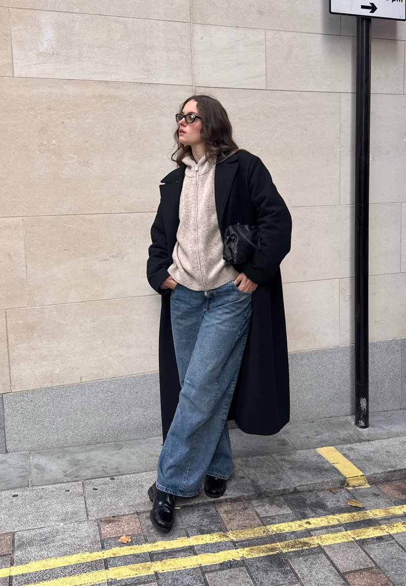 Cappotto lungo nero sopra un maglione beige a maglia, jeans blu a gamba larga e scarpe nere. Gli accessori includono una piccola borsa nera. Sfondo urbano.