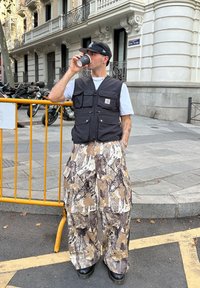 Muž v čiernej čiapke, čiernej veste a širokých maskáčových nohaviciach pije z čierneho pohára pri žltom zábradlí na ulici.