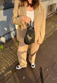 Costume beige ajusté avec un blazer à un seul bouton et un pantalon ample, associé à un débardeur blanc côtelé et un sac à cordon en cuir noir.