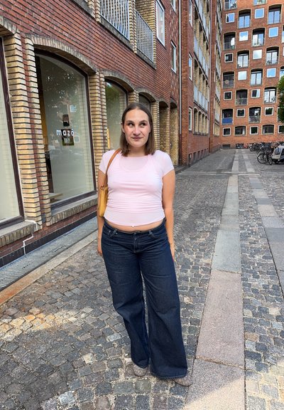 Mujer con una blusa rosa y vaqueros anchos de pierna oscura, de pie en una calle empedrada bordeada de edificios de ladrillo y bicicletas.