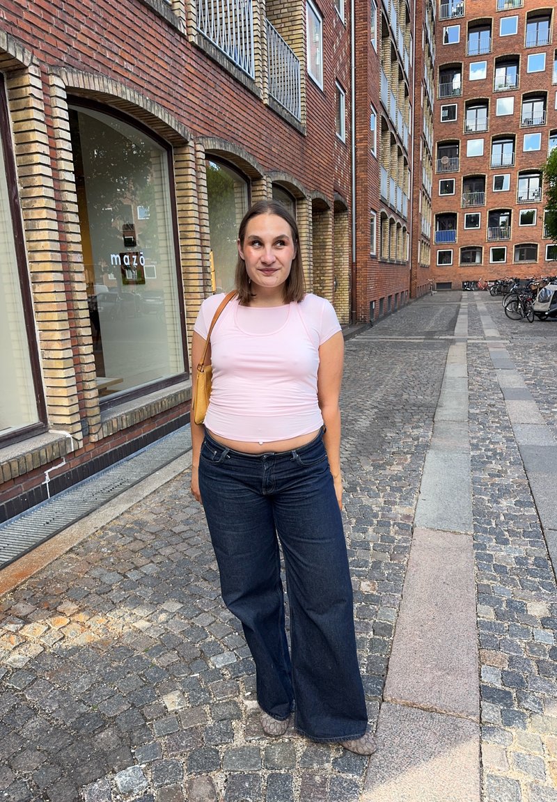 Femme portant un haut rose et un jean large sombre, debout dans une rue pavée bordée de bâtiments en briques et de vélos.