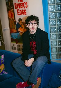 Young man with glasses and moustache sits on blue sofa wearing black blazer, pinstripe trousers, red trainers, with movie poster in the background.
