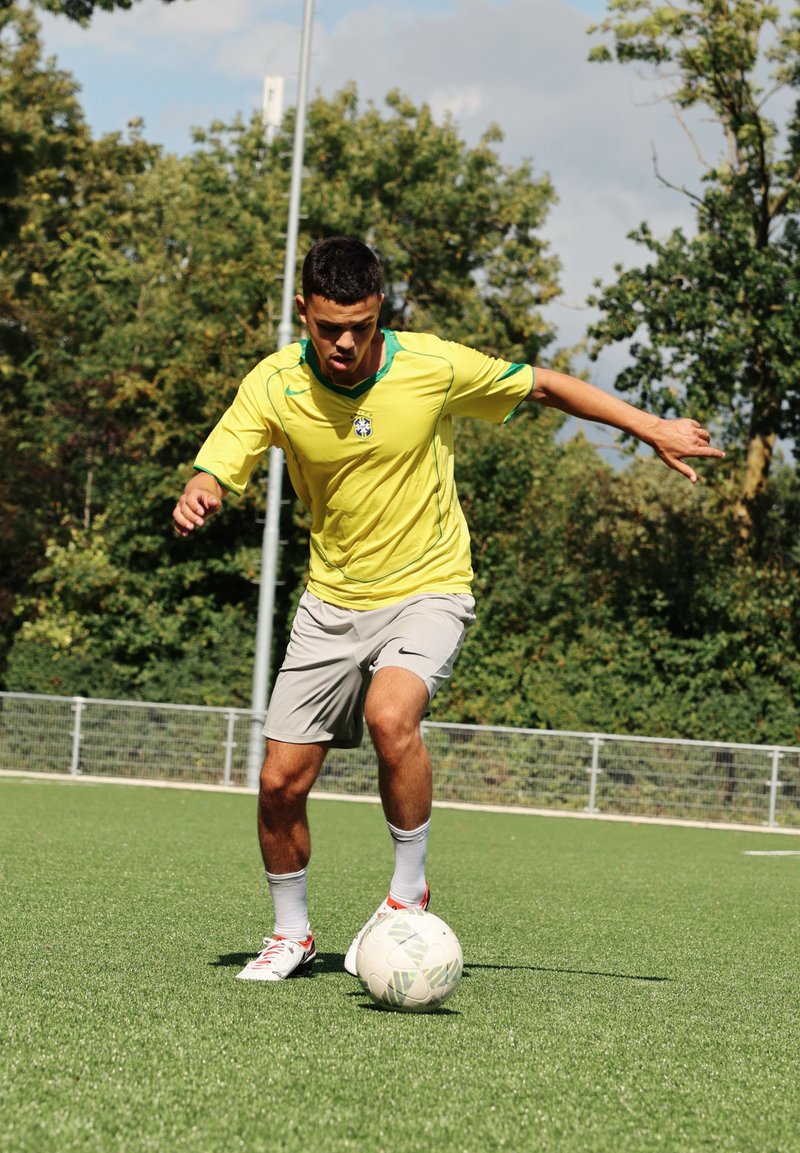 Atleta com uma camisola brilhante amarela e verde e calções cinza, a driblar uma bola de futebol branca num campo de relva verde.