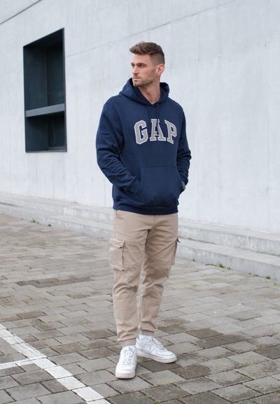 Joven con sudadera azul marino con el logo de GAP, pantalones cargo beige y zapatillas blancas, de pie sobre un suelo pavimentado cerca de una pared de concreto gris.