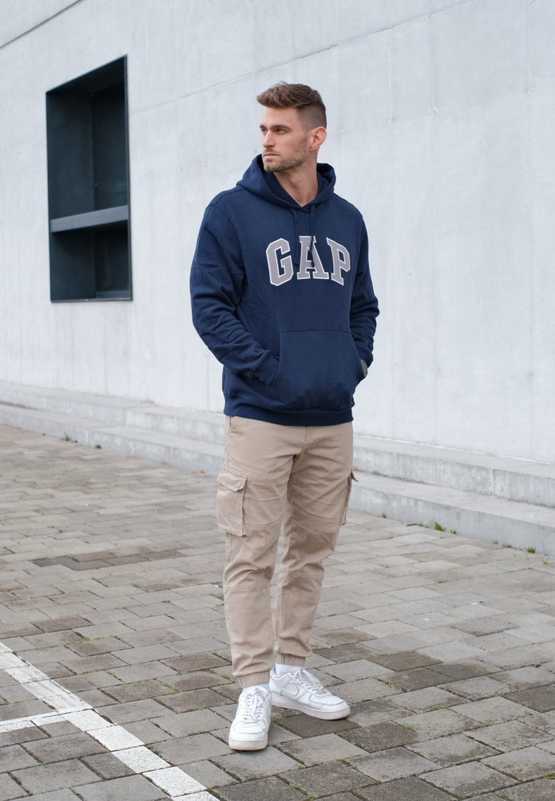 Jeune homme portant un sweat à capuche bleu marine avec le logo GAP, un pantalon cargo beige et des baskets blanches, debout sur un sol pavé près d'un mur en béton gris.
